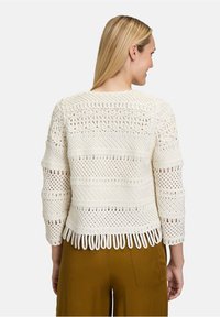 Pull en crochet couleur crème avec des motifs ajourés complexes, une texture côtelée et des franges au bas. Manches trois-quarts et col rond.