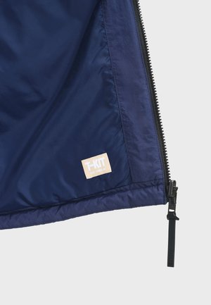Interiér námořnicky modré bundy s viditelným zavíráním na zip napravo a béžovou štítkem s nápisem "T-KIT TOUT TEMPS UR."