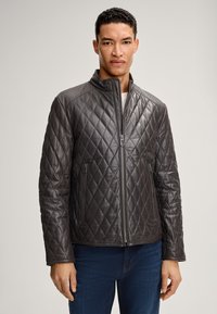 Mann in einer dunkelgrauen, gesteppte Lederjacke mit Reißverschluss und hohem Kragen sowie blauen Jeans, der vor einem einfarbigen Hintergrund steht.