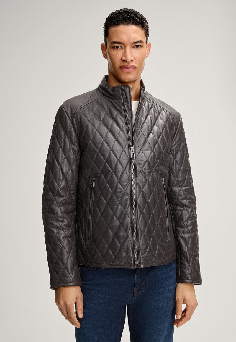 Mann in einer dunkelgrauen, gesteppte Lederjacke mit Reißverschluss und hohem Kragen sowie blauen Jeans, der vor einem einfarbigen Hintergrund steht.