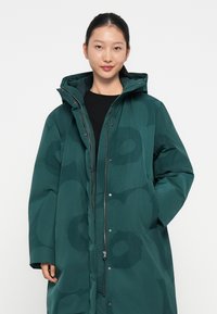 Zils kapuci parka ar spīdīgu apdari, ar smalku rakstu, priekšējo rāvējslēdzēju un pogām, kas izstrādāta siltumam un komfortam.