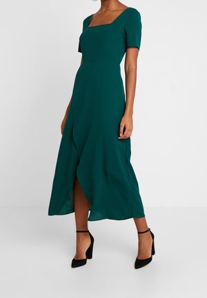 Femme portant une robe midi verte à manches courtes et encolure carrée, associée à des talons hauts noirs à bride à la cheville, debout contre un mur blanc.