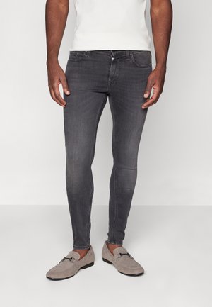 Mann trägt schlanke, ausgewaschene schwarze Jeans und hellbraune Wildleder-Slipper, steht vor hellem Hintergrund.