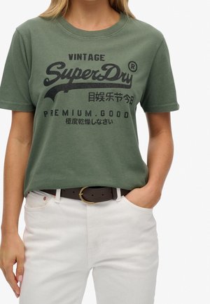 Frau trägt grünes SuperDry Vintage-Logo-T-Shirt, in weiße Hose mit schwarzem Gürtel gesteckt, Hand in der Tasche, beschnitten an Hals und Oberschenkeln.