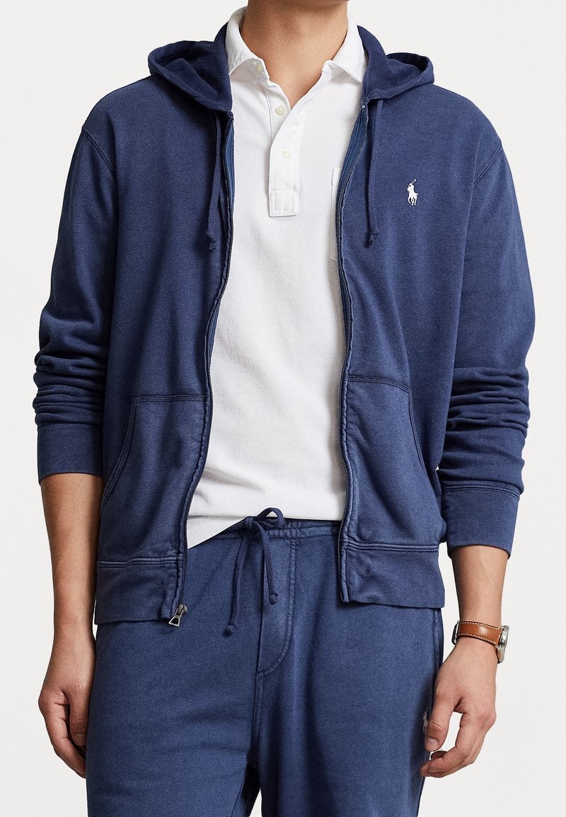 Mörkblå zip-up hoodie i mjukt tyg med en framficka, ribbade ärmslut och huva med dragsko. Kombinerad med en vit polo under.