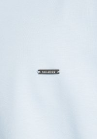 T-shirt azzurro chiaro realizzato in tessuto morbido, con un piccolo tassello metallico argentato riportante la scritta "SMILOBX" inciso, posizionato sulla parte anteriore.