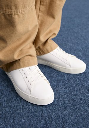 Pieds portant des baskets blanches à lacets et un pantalon beige, debout sur un sol recouvert d'une moquette bleue.