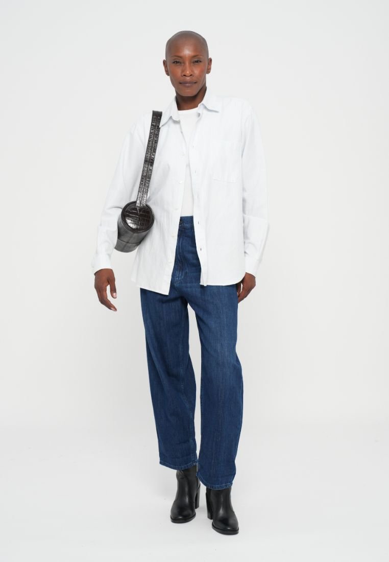 Chemise blanche à boutons, pantalon en denim bleu, bottes à talons noires et sac rond noir. La chemise a une coupe décontractée avec deux poches poitrine.