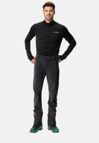 Sudadera térmica negra con cremallera y logo, y pantalones ajustados negros. Botas de esquí verdes. Presenta un diseño ajustado y una textura de tela suave.