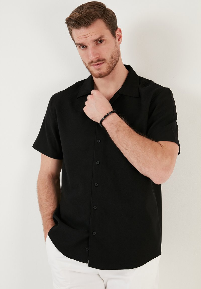 Zwarte korte- mouw button-up shirt met een campingkraag, zachte textuur en een relaxte pasvorm, gecombineerd met witte shorts en een zwarte armband.