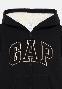 Casaco com fecho éclair preto, forrado a fleece branco e grande logotipo GAP contornado na parte da frente, tamanho 10 crianças.