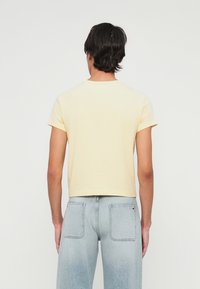 Persona con cabello corto y oscuro, vistiendo una camiseta lisa de color amarillo claro y jeans azules claros, de pie de espaldas contra un fondo blanco.