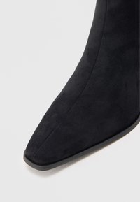 Bota de tornozelo em camurça preta com bico fino e textura suave, apresentando um salto baixo preto e uma silhueta elegante.