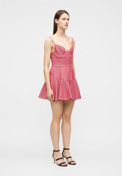 Femme debout portant une mini robe en denim rose sans manches avec des bretelles fines et des sandales noires à talons hauts, sur un fond blanc uni.