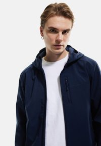 Chaqueta con capucha de color navy, de textura suave y con un bolsillo frontal con cremallera. Se lleva encima de una camiseta blanca de cuello redondo, que presenta un diseño ajustado.