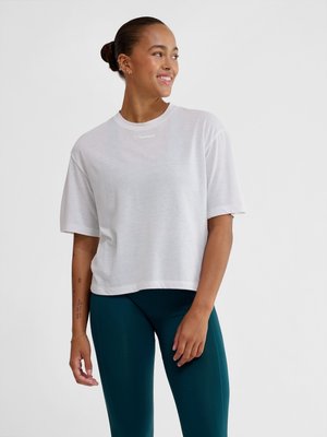 Kobieta uśmiechająca się i patrząca w prawo, ubrana w luźny biały krótki rękaw i ciemnoturkusowe legginsy, stoi na tle jednolitego jasnego tła.