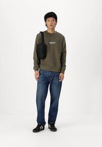 Sudadera verde oliva con texto blanco, jeans azules y zapatillas negras. Pequeña bolsa de hombro negra cuelga del hombro. Fondo sencillo.