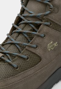 Brown hiking shoe featuring textured fabric, suede panels, blue laces, and a small embossed logo. Curved design with reinforced stitching.