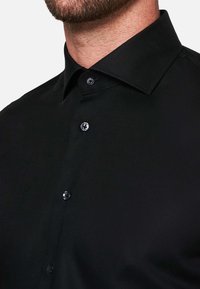 Zwart overhemd met knoopsluiting, gestructureerde kraag, twee knopen aan de voorkant en een gladde textuur. De stof lijkt lichtgewicht en strak.