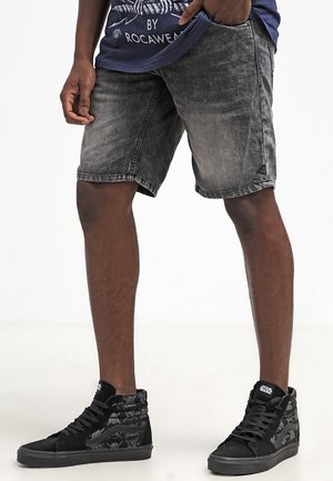 Schwarze Denim-Shorts mit verwaschenem Finish, geradem Schnitt und seitlichen Taschen, kombiniert mit schwarzen High-Top-Sneakern mit Muster.