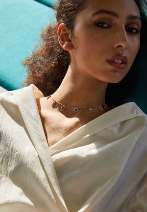 Nuori nainen, jolla on kiharat hiukset, valkoinen paita ja hienovarainen kultainen kaulakoru, jossa on kirjaimen ja silmän muotoisia riipuksia.