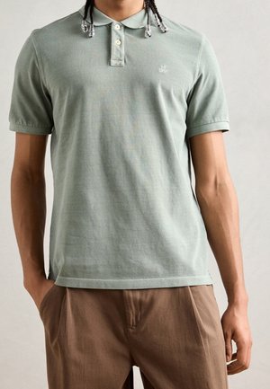 Mann trägt hellgrünes kurzärmeliges Polohemd und braune Hose mit Händen in den Taschen vor einfachem Hintergrund.