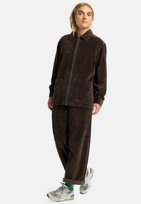Brown corduroy jacket with a zip front, large pockets, and a collar, paired with matching corduroy pants and gray sneakers.
