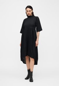 Sieviete ar tumšiem mitriem matiem, tērpusies melnā vidēja garuma kleitā ar augstu apkakli un elkoņa garuma piedurknēm, kājās melni potīšu zābaki, stāvot pret baltu vienmuļu fonu.