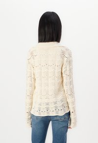 Strickpullover in Creme, mit einem offenen Muster mit Wellen-Designs. Lange Ärmel und einen abgerundeten Saum. Kombiniert mit blauen Jeans.