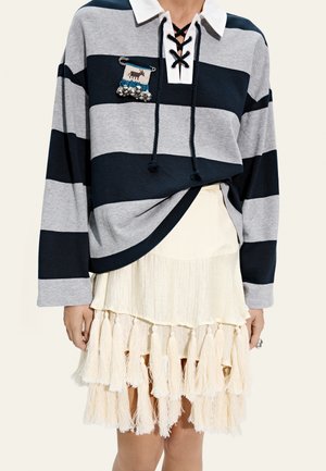 Kobieta ubrana w czarną i szarą pasiastą koszulę z długim rękawem, białym kołnierzykiem i kremową spódnicę z warstwami frędzli na dole.