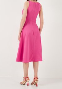 Robe sans manches fuchsia avec un bodice ajusté et une jupe évasée, présentant une fermeture éclair au dos et une texture lisse ; portée avec des sandales à talons.