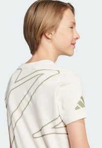 Camiseta de algodón de manga corta en color crema, con patrones de líneas abstractas verdes en la parte posterior y un logo de Adidas en la manga.