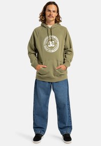 Felpa verde oliva con un grande logo bianco, tasca a kangaroo e cappuccio con cordino. Abbinata a jeans blu larghi e sneakers nere.