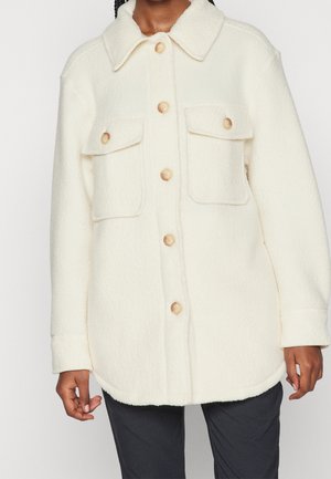 Roomkleurig fleecejack met knopen en twee borstzakken met klep, gedragen over zwarte broek.