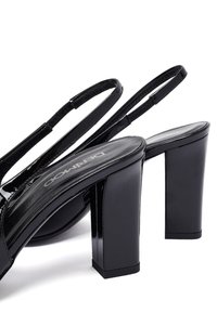 Sandalias de charol negro con tiras al tobillo, tacones de bloque y puntera cuadrada sobre un fondo blanco.