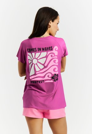 Vrouw met lang donker haar die een paarse t-shirt draagt met roze golven, bloemmotieven en de tekst "COMES IN WAVES PROTEST" op de rug.