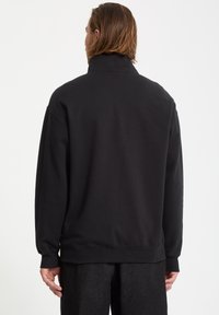 Felpa nera con colletto alto, vestibilità rilassata e polsini a coste. La texture sembra morbida; cuciture dettagliate visibili lungo le cuciture.