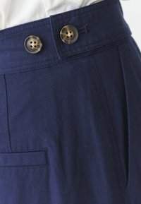 Pantalones azul marino con cierre de doble botón, diseño con pliegues y un bolsillo lateral con un acabado de tejido texturizado.