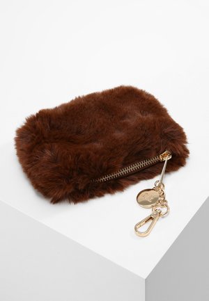 Pochette en fausse fourrure marron avec fermeture éclair dorée, breloque ronde et mousqueton, posée sur une surface blanche inclinée.