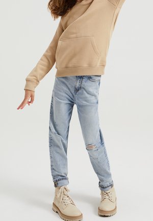 Sudadera beige clara con un bolsillo delantero, combinada con jeans mom de lavado claro que tienen rotos, y botas beige con cordones y suela texturizada.