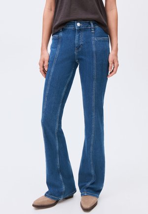 Vrouw in hoge blauwe denim jeans met een voornaaddetail enbeige puntige schoenen, staande tegen een effen achtergrond.
