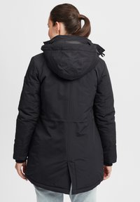 Parka sombre et isolée avec capuche, présentant une longueur au dos, des poignets élastiques et une finition lisse et mate.