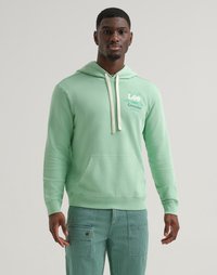 Uomo che indossa un felpa verde chiaro con i loghi Lee e Crayola, pantaloni cargo verde chiaro, in piedi su uno sfondo semplice.