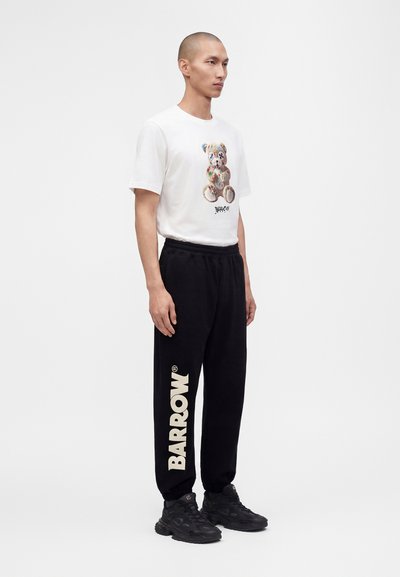 Homme portant un t-shirt blanc avec un motif coloré d'ours en peluche et un pantalon de jogging noir avec un grand texte "Barrow" sur la jambe gauche, debout sur fond blanc.
