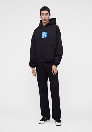 Joven vestido con una sudadera negra con capucha con el logo azul de "Karl Lagerfeld Jeans", pantalones negros y zapatillas negras y blancas, de pie sobre un fondo blanco.