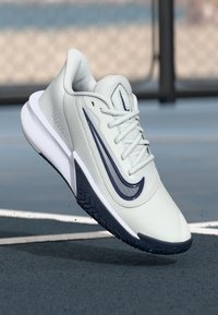 White and navy athletic shoe with laces, raised sole, and textured grip, positioned as if mid-air on a sports court background.