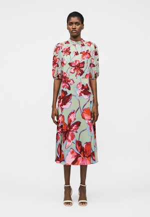 Γυναίκα όρθια, φορόντας ένα ανοιχτοπράσινο midi φόρεμα με μεγάλα κόκκινα και ροζ λουλουδένια σχέδια και κοντά φουσκωτά μανίκια, σε συνδυασμό με λευκές σανδάλες με τακούνι.