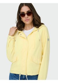 Femme portant des lunettes de soleil œil-de-chat et une veste à capuche jaune clair avec des poches avant, ainsi qu'un jean bleu, debout devant un fond uni.