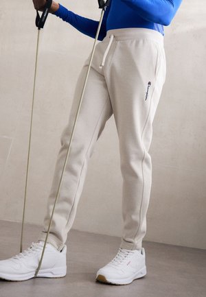 Persona con pantalones deportivos beige de Champion y zapatillas blancas usando bandas de resistencia para hacer ejercicio en interiores.