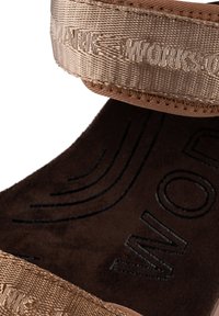 Ruskea sandaali, jossa on teksturoitu kangasremmi, kohokuvioitu logo ja pehmeä mokkasiinin sisäpohja. Ominaisuuksiin kuuluu litteä pohja ja monimutkainen ompeluseikka.